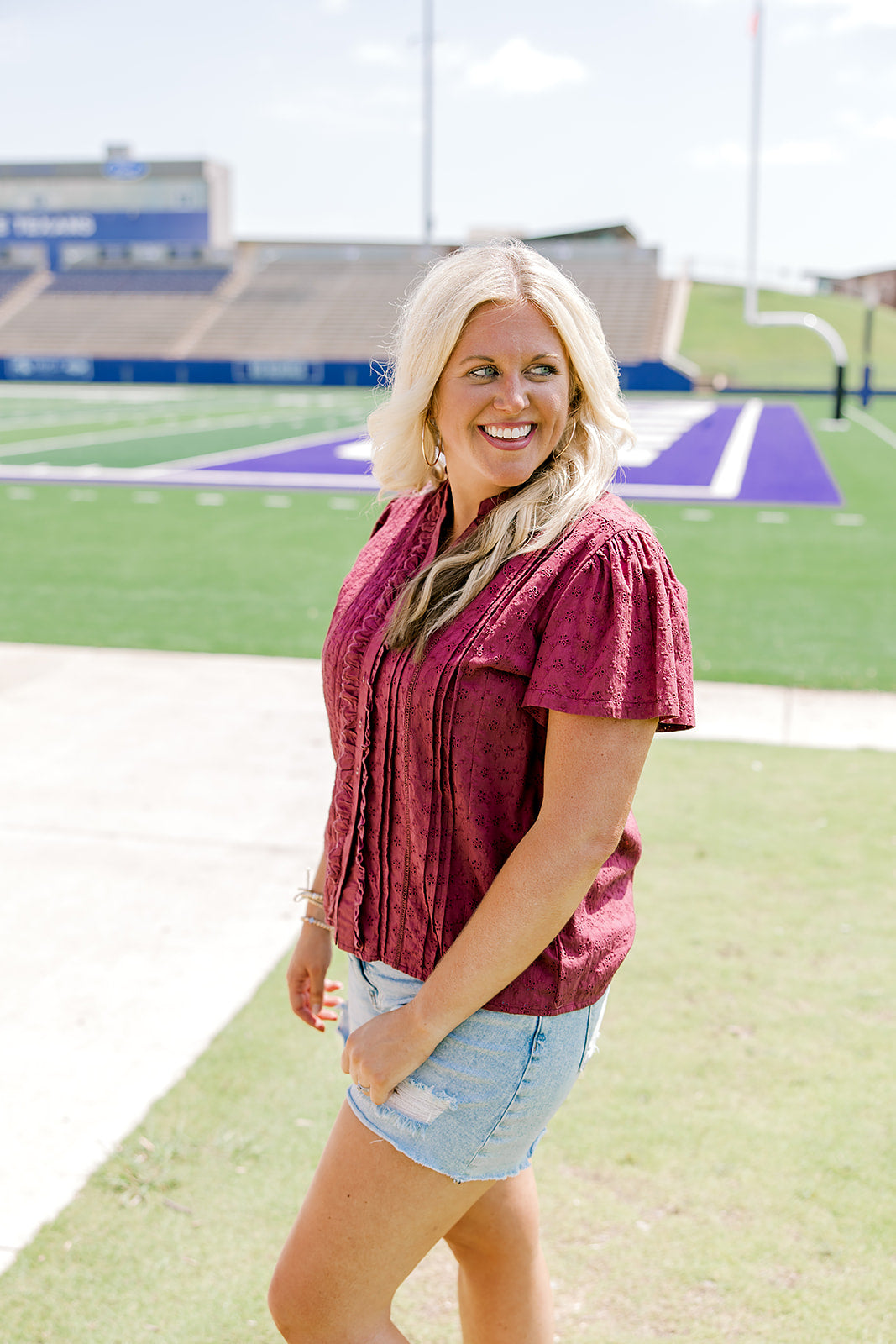 Woman with blonde hair standing in a side profile position wearing the Lilly Top in a lifestyle scene. The focal point of this image is the pleating on sleeve as well as maroon eyelet pattern across top. The model paired this top with light-wash denim shorts. 