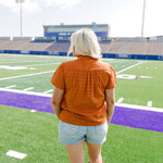 Woman with blonde hair standing in a back profile position wearing a short-sleeve top that's burnt orange eyelet pattern, and light-wash denim shorts. 