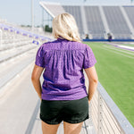 Woman with blonde hair standing in a back profile position wearing the Lilly Top in a lifestyle scene. The focal point of this image is the pleating on back of top and purple eyelet pattern. The model paired this top with black denim shorts. 