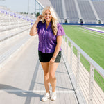 Woman with blonde hair standing in a front profile position wearing the Lilly Top in a lifestyle scene. The focal point of this image is the ruffles along front of top, v-neckline, and purple eyelet pattern. The model paired this top with black denim shorts. 