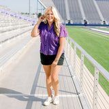 Woman with blonde hair standing in a front profile position wearing the Lilly Top in a lifestyle scene. The focal point of this image is the ruffles along front of top, v-neckline, and purple eyelet pattern. The model paired this top with black denim shorts. 