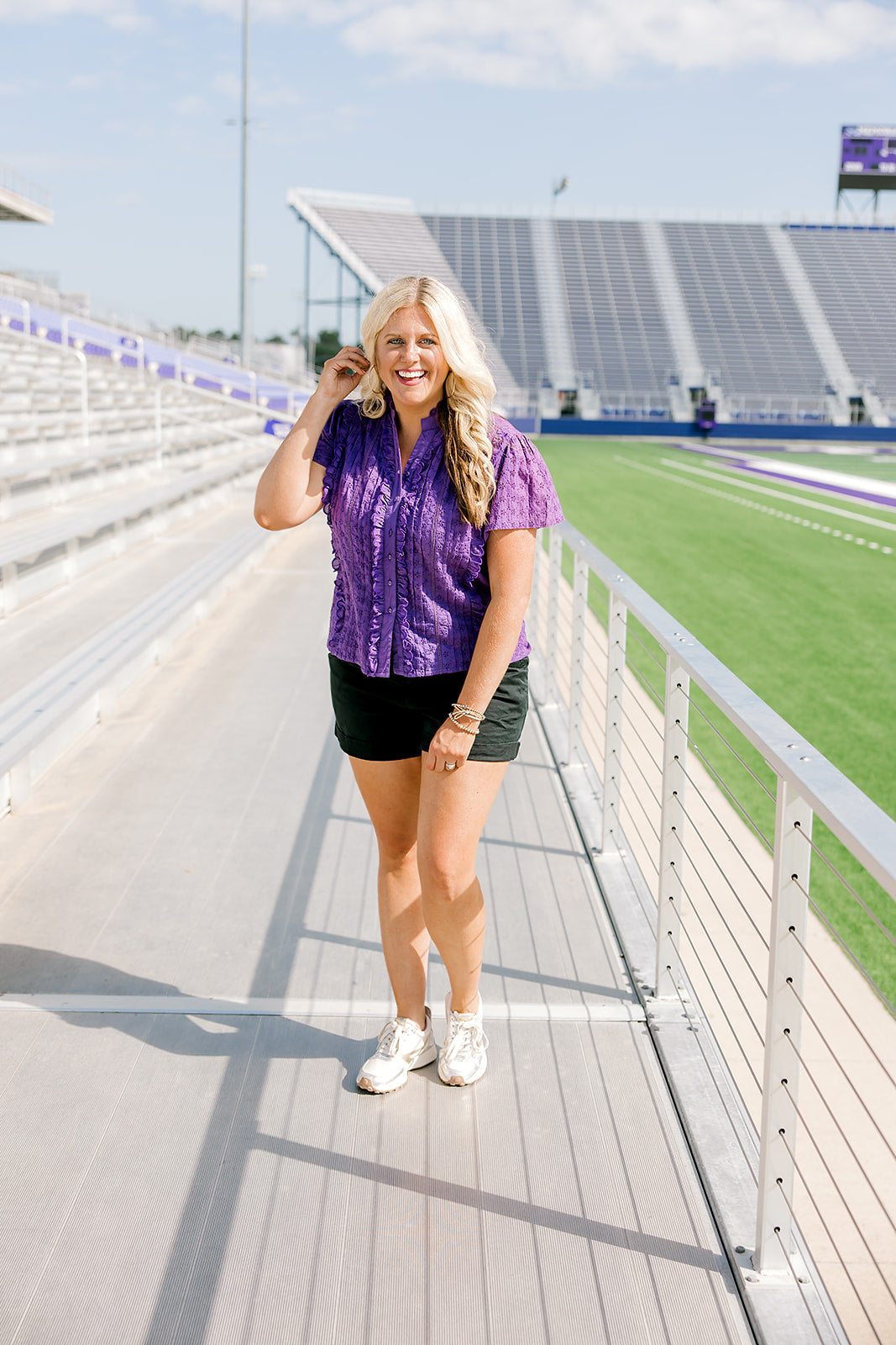 Woman with blonde hair standing in a front profile position wearing the Lilly Top in a lifestyle scene. The focal point of this image is the ruffles along front of top, v-neckline, and purple eyelet pattern. The model paired this top with black denim shorts. 