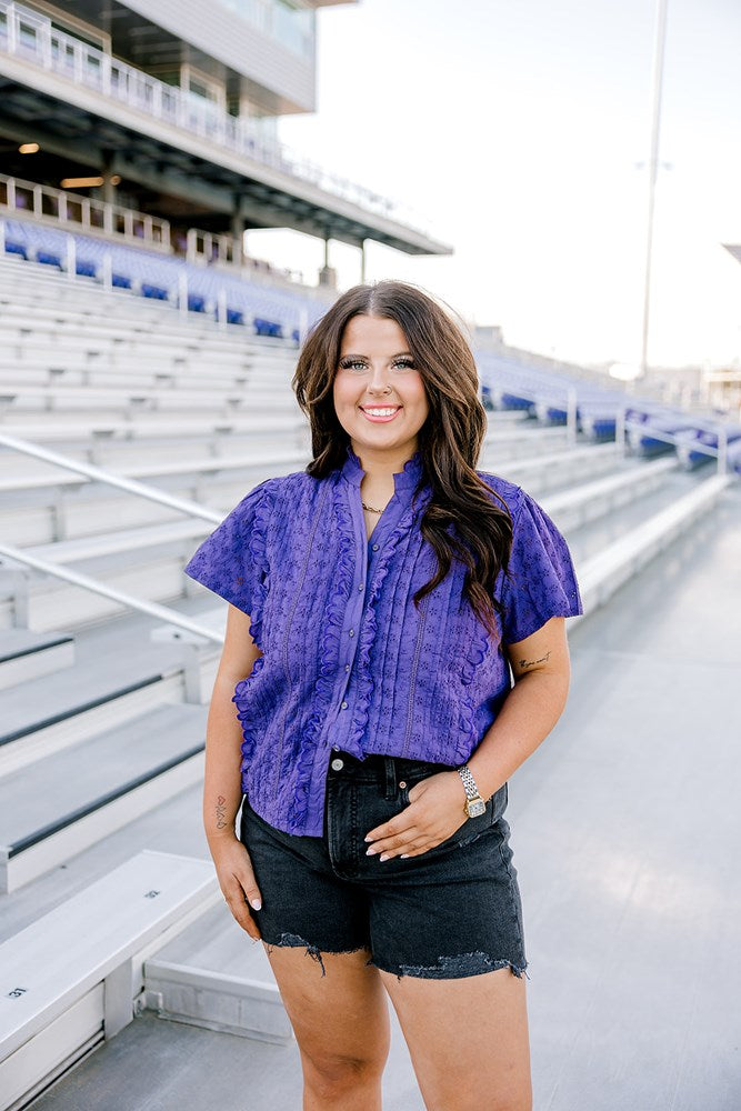 Woman with dark hair standing in a front profile position wearing the Lilly Top in the stands of a football stadium. The focal point of this image is the purple eyelet pattern, ruffles down side of buttons and collar. The model paired this top with black denim shorts.