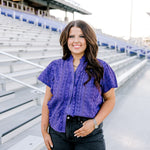 Woman with dark hair standing in a front profile position wearing the Lilly Top in the stands of a football stadium. The focal point of this image is the purple eyelet pattern, ruffles down side of buttons and collar. The model paired this top with black denim shorts.