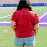 Woman facing backwards wearing a short-sleeve red eyelet top and light-wash denims shorts. 