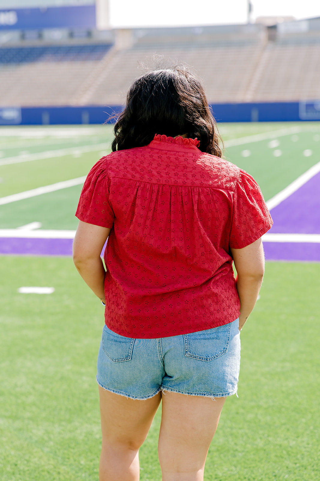 Woman facing backwards wearing a short-sleeve red eyelet top and light-wash denims shorts. 