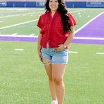 Model facing to the side wearing a short-sleeve red top with eyelet pattern and light-wash denim shorts. 