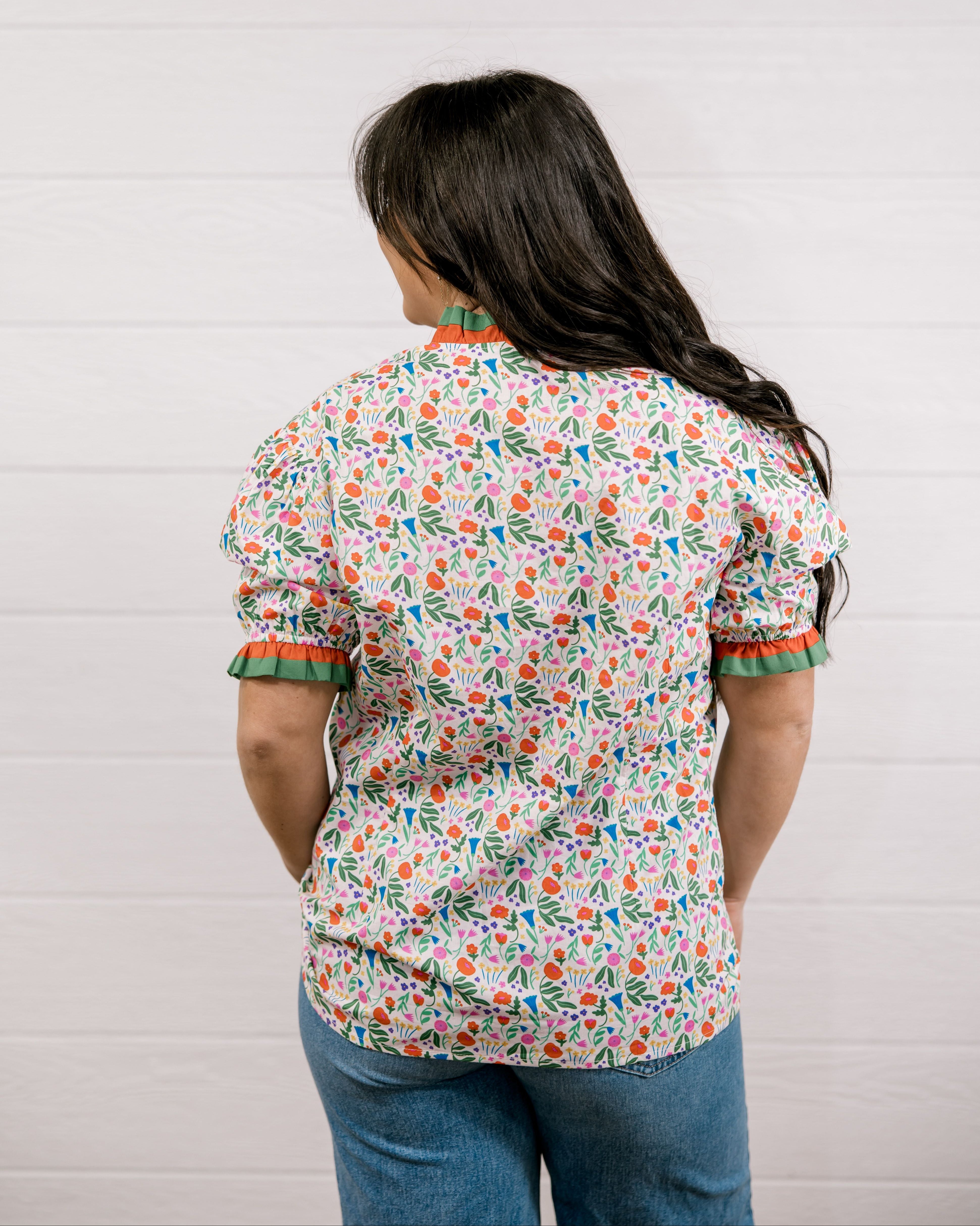 A women wearing a floral top with green and red trim with a ruffle collar and sleeves showing the back