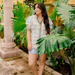 A women wearing a floral top with green and red trim with a ruffle collar and sleeves in a natural stone atmosphere 
