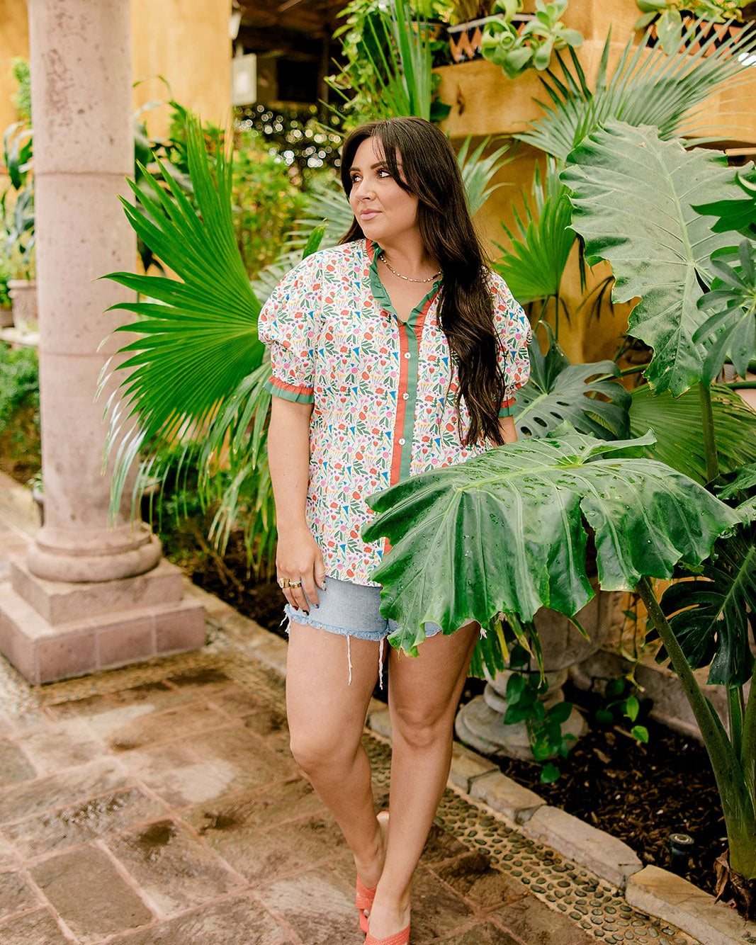 A women wearing a floral top with green and red trim with a ruffle collar and sleeves in a natural stone atmosphere 