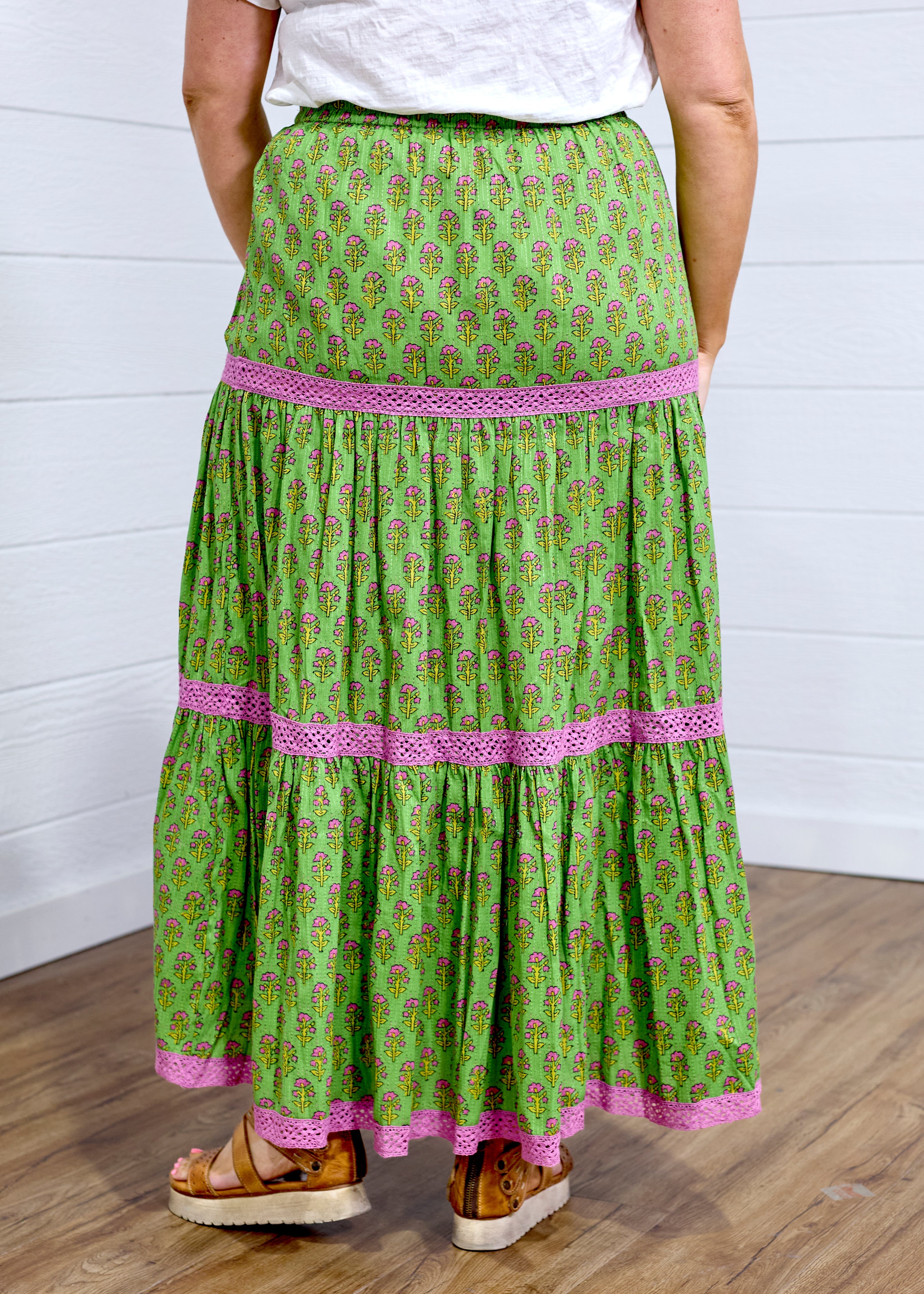 Model in a back profile position against a white background in the Madison Maxi Skirt. The image portrays length of skirt, the embroidery, and graphics on back of skirt. Model paired this with brown sandals to elevate look.