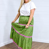 Woman with blonde hair in front facing position wearing the Madison Maxi Skirt against a white background. The images focal point is on the front of the skirt showing it's length, embroidery, and front graphics. Model paired skirt with white top and brown sandals. 