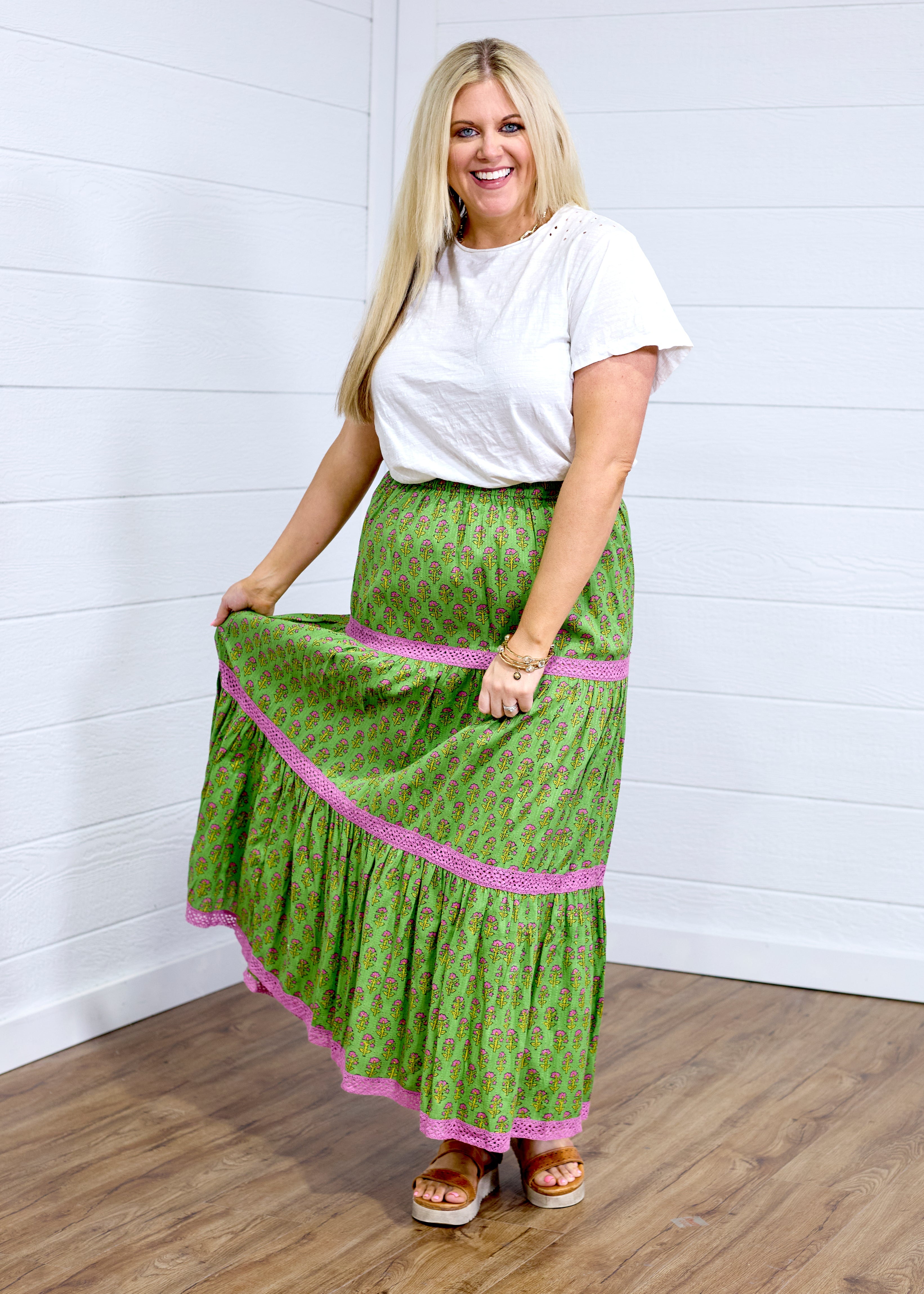 Woman with blonde hair in front facing position wearing the Madison Maxi Skirt against a white background. The images focal point is on the front of the skirt showing it's length, embroidery, and front graphics. Model paired skirt with white top and brown sandals. 