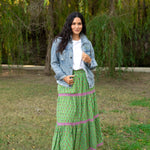 Model with dark hair in a lifestyle scene wearing the Madison Maxi Skirt. The background of this image is heavy in greenery. Model paired skirts with white top, denim jacket, and white sandals. 