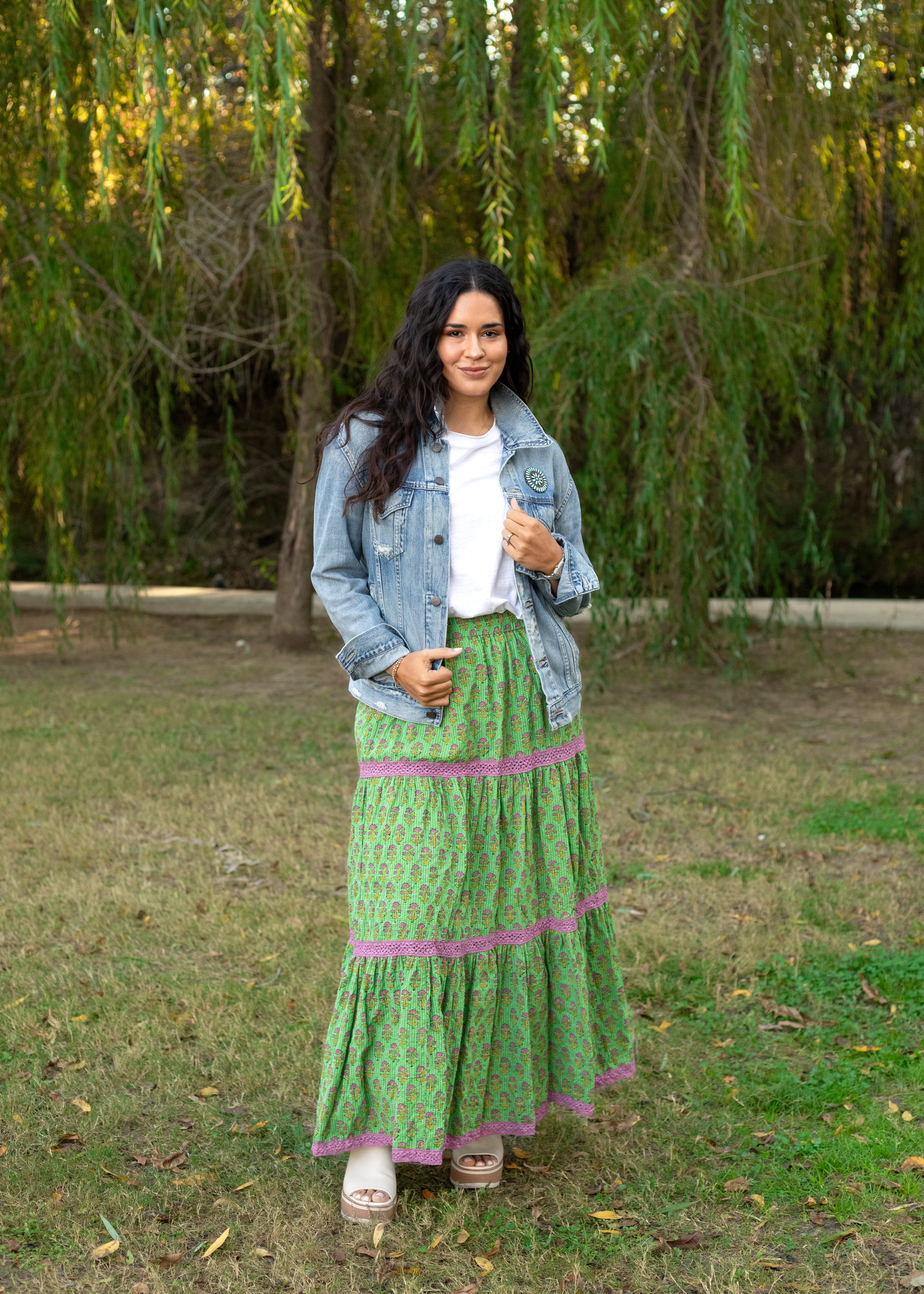 Model with dark hair in a lifestyle scene wearing the Madison Maxi Skirt. The background of this image is heavy in greenery. Model paired skirts with white top, denim jacket, and white sandals. 