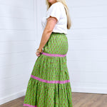 Woman with blonde hair in side profile position wearing the Madison Maxi Skirt against a white background. The images focal point is the side of skirt, expressing it's length and embroidery embellishments on the sides. Model paired with white top and brown sandals.