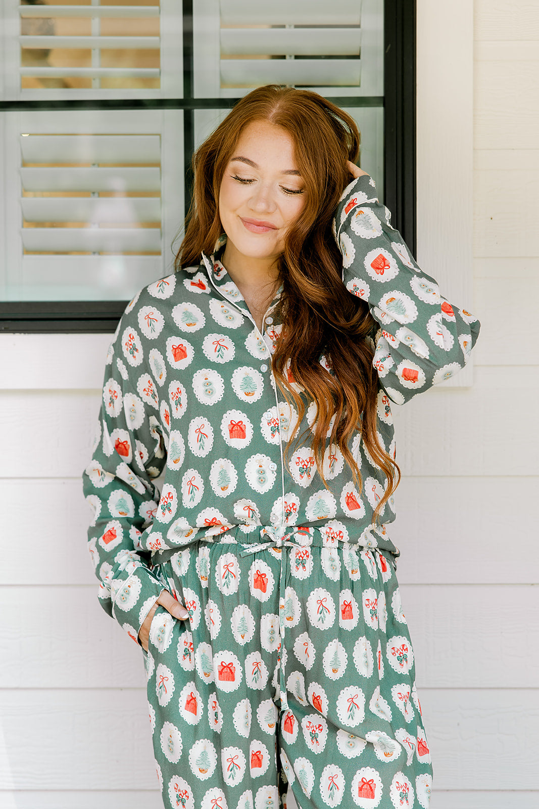 Woman with red hair wearing a long-sleeve top and long pant pajama set. The Pajamas feature fun red & green Christmas icon graphics. 