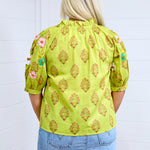 Woman with blonde hair facing in a back profile against a white back ground. The image shows embroidered flowers on back of sleeves and graphics on back. She has it paired with a light-wash denim skirt. 