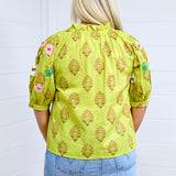 Woman with blonde hair facing in a back profile against a white back ground. The image shows embroidered flowers on back of sleeves and graphics on back. She has it paired with a light-wash denim skirt. 