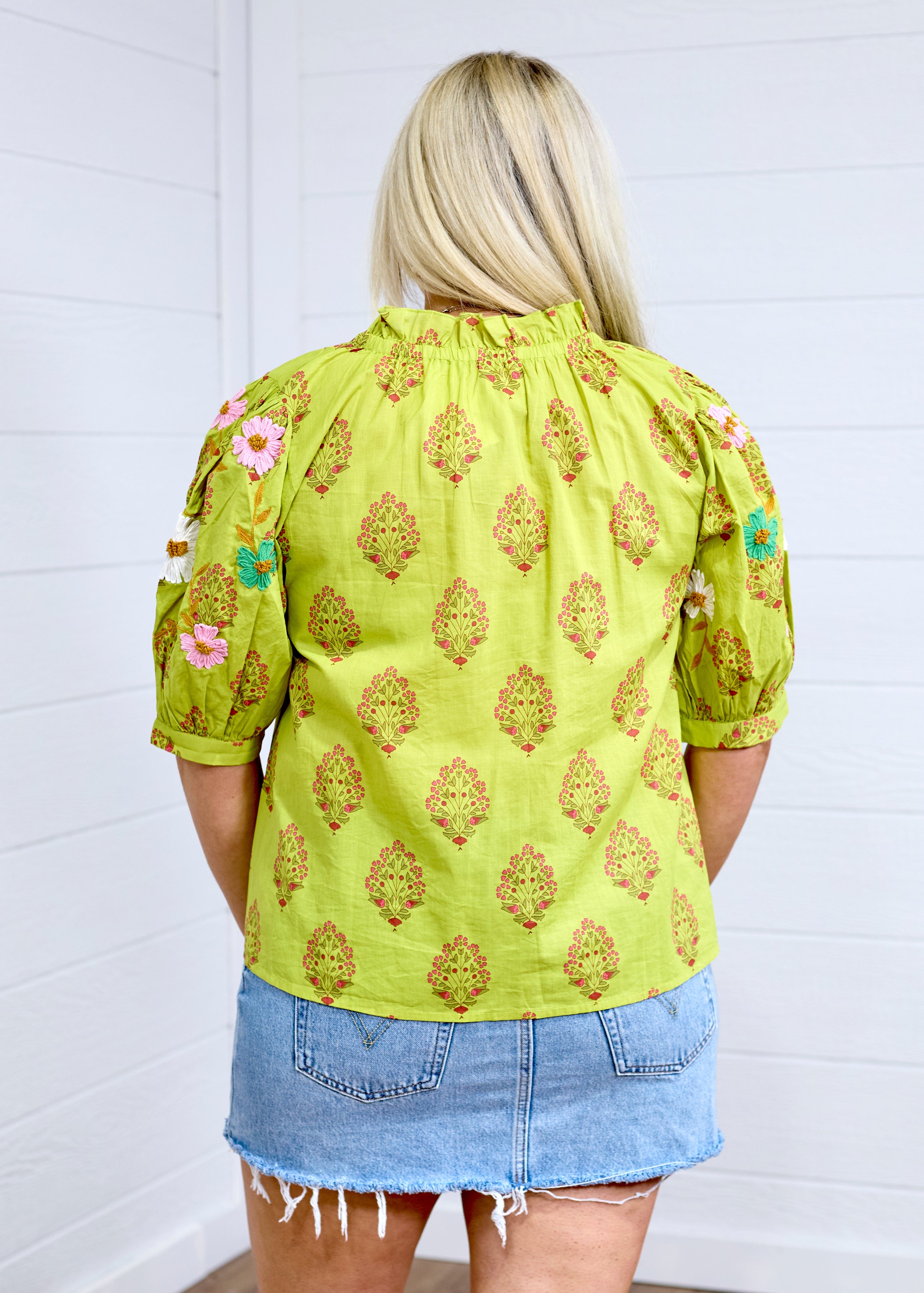 Woman with blonde hair facing in a back profile against a white back ground. The image shows embroidered flowers on back of sleeves and graphics on back. She has it paired with a light-wash denim skirt. 