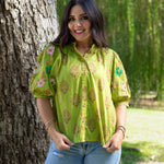 Woman with dark hair showing front profile of the Margo Top in a lifestyle scene. The back ground is wooded and hints of heavy greenery. She has the top paired with light-wash jeans. 