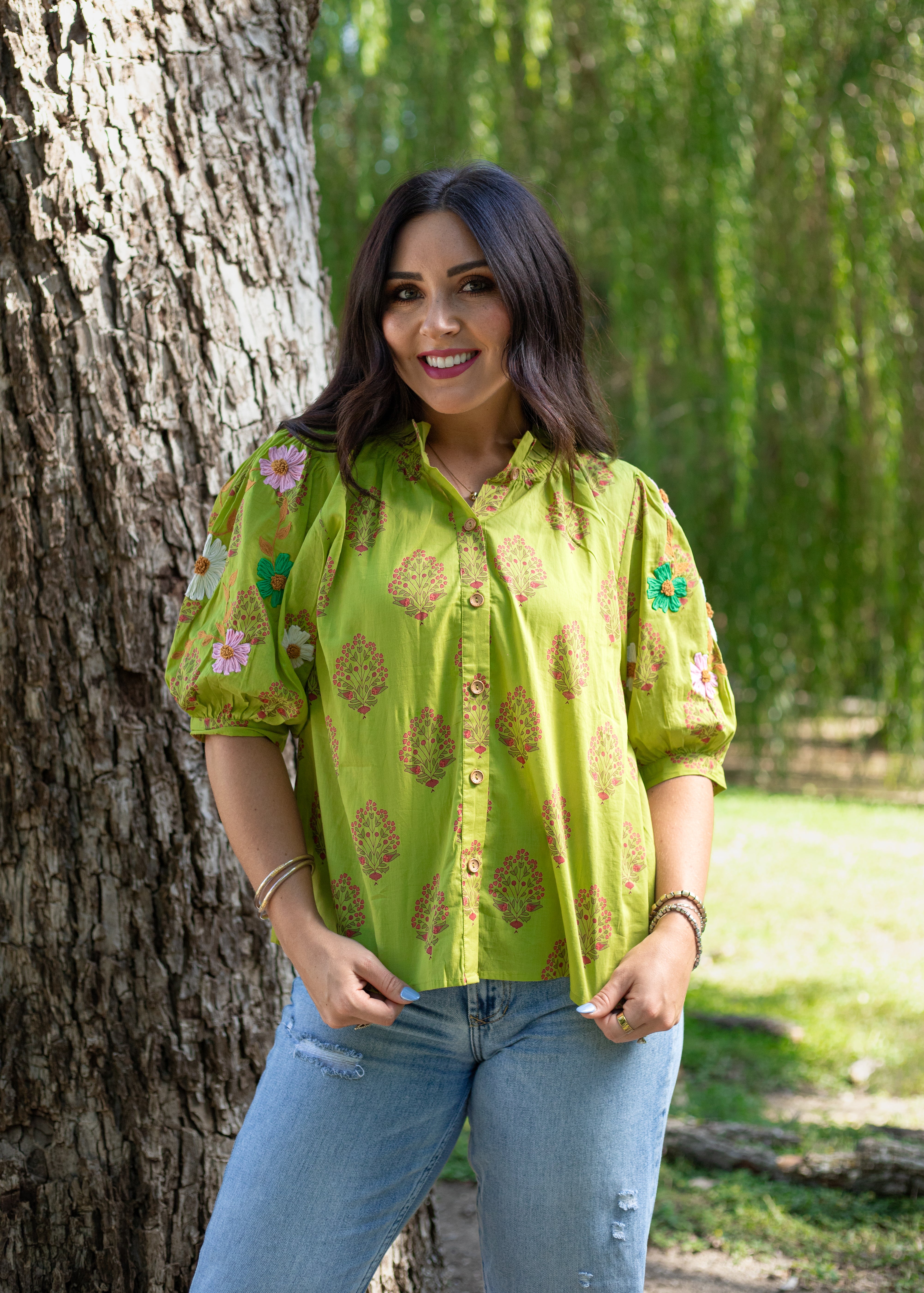 Woman with dark hair showing front profile of the Margo Top in a lifestyle scene. The back ground is wooded and hints of heavy greenery. She has the top paired with light-wash jeans. 