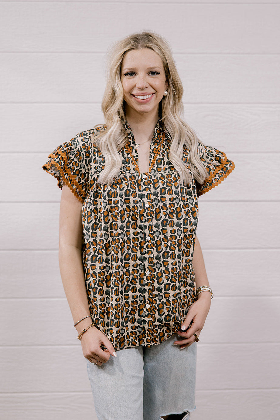 A woman with blonde hair wearing a brown and beige leopard print short-sleeve blouse with ruffled trim, paired with light-wash ripped jeans, standing against a white wood-paneled wall.