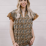 A woman with blonde hair wearing a brown and beige leopard print short-sleeve blouse with ruffled trim, paired with light-wash ripped jeans, standing against a white wood-paneled wall.