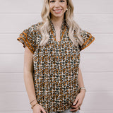 A woman with blonde hair wearing a brown and beige leopard print short-sleeve blouse with ruffled trim, paired with light-wash ripped jeans, standing against a white wood-paneled wall.