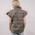 A woman viewed from behind wearing a short-sleeve button-up shirt with a brown, black, and beige animal print and orange patterned trim on the sleeves, paired with light blue jeans against a white wood-paneled wall.