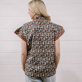 A woman viewed from behind wearing a short-sleeve button-up shirt with a brown, black, and beige animal print and orange patterned trim on the sleeves, paired with light blue jeans against a white wood-paneled wall.