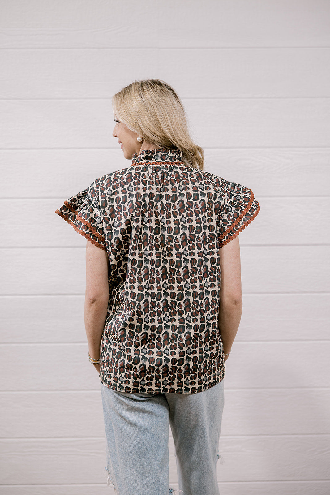 A woman viewed from behind wearing a short-sleeve button-up shirt with a brown, black, and beige animal print and orange patterned trim on the sleeves, paired with light blue jeans against a white wood-paneled wall.