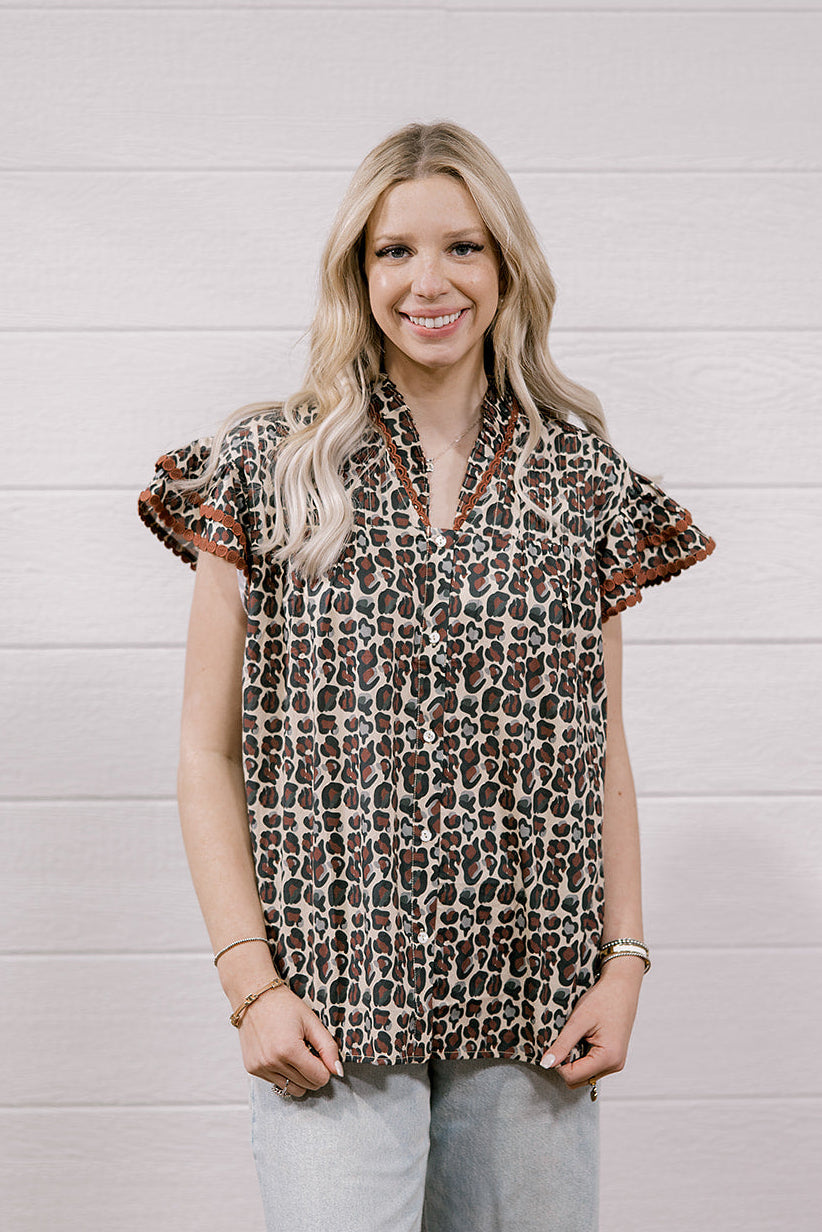 A woman modeling a brown and beige leopard print short-sleeve blouse with ruffled sleeves, paired with light-wash ripped jeans, standing against a white wood-paneled wall.