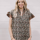 A woman modeling a brown and beige leopard print short-sleeve blouse with ruffled sleeves, paired with light-wash ripped jeans, standing against a white wood-paneled wall.