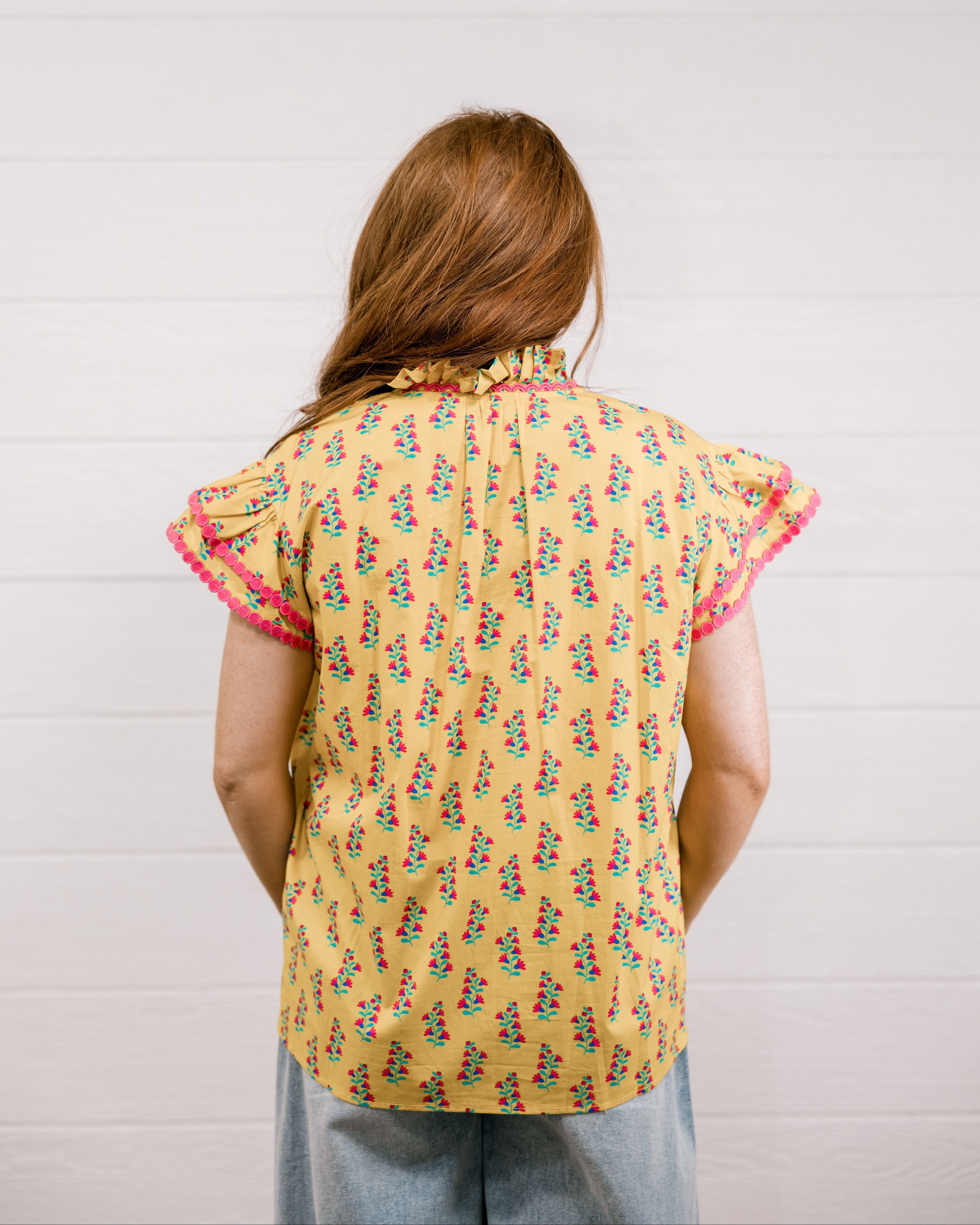 Women wearing a yellow top with floral print with ruffle sleeves and pink trim showing the back on a white background 
