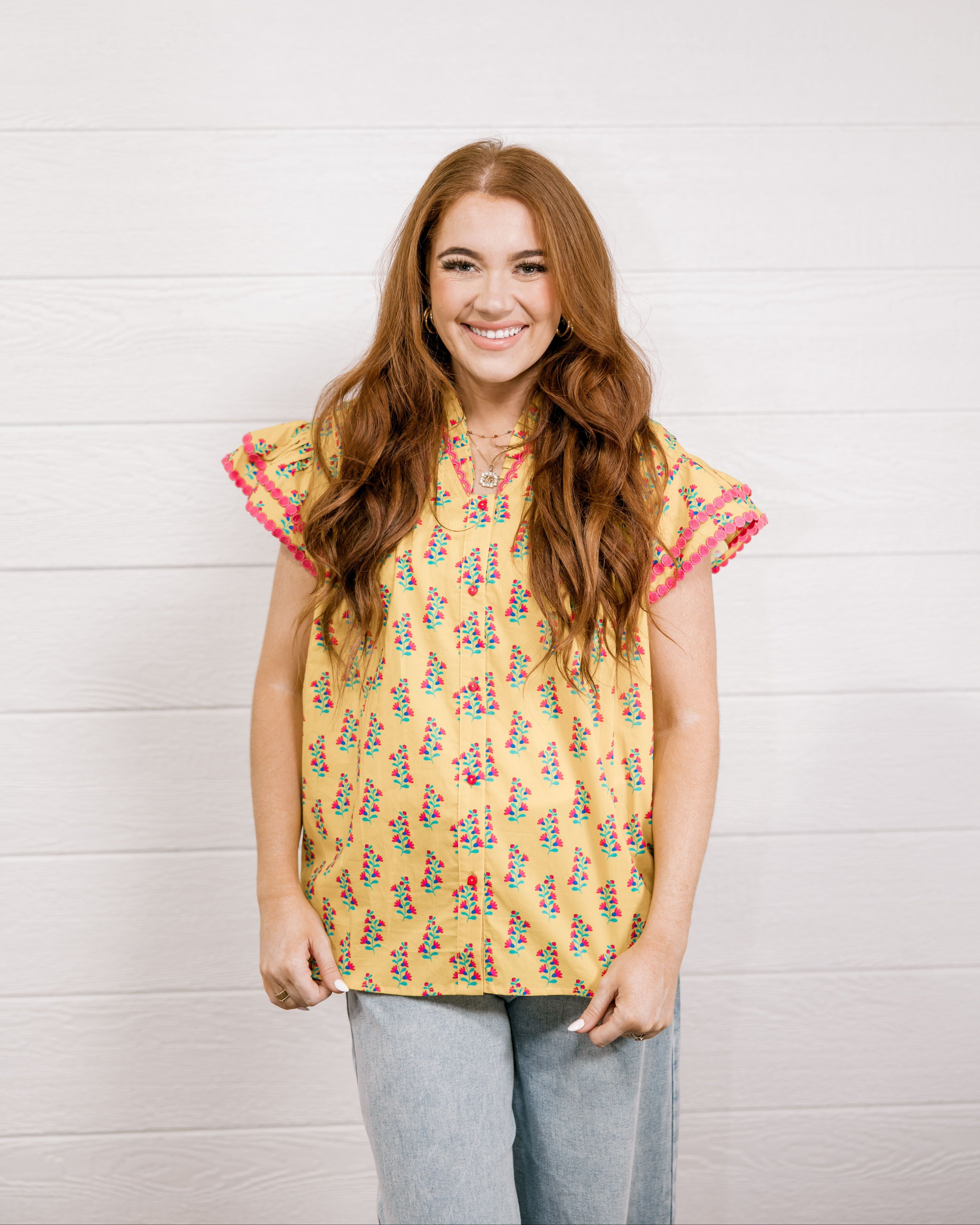 Women wearing a yellow top with floral print with ruffle sleeves and pink trim showing the front on a white background 