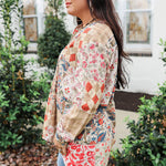 Woman with dark hair standing in a side profile position wearing the Nina Top in a lifestyle scene. The focal point of this image is the red/blue patchwork print and well as the long sleeves and length of top. The model paired this top with light-wash denim jeans. 