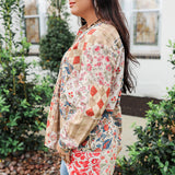 Woman with dark hair standing in a side profile position wearing the Nina Top in a lifestyle scene. The focal point of this image is the red/blue patchwork print and well as the long sleeves and length of top. The model paired this top with light-wash denim jeans. 