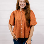 A woman with long light brown hair wearing a rust-orange short-sleeve blouse with white buttons and dark blue jeans, standing against a white wood-paneled wall.