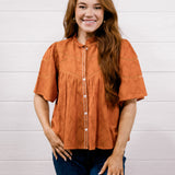 A woman with long light brown hair wearing a rust-orange short-sleeve blouse with white buttons and dark blue jeans, standing against a white wood-paneled wall.