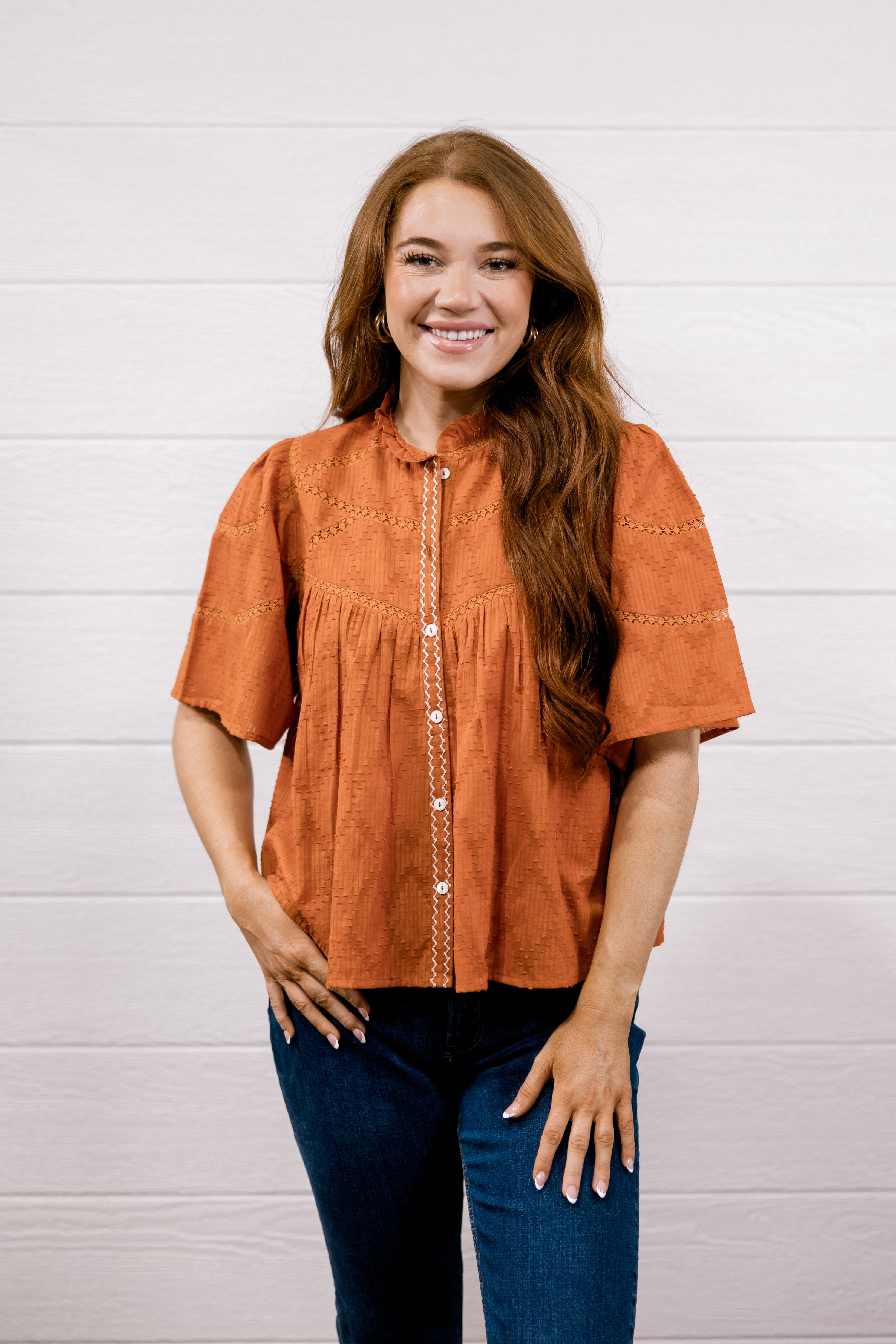 A woman with long light brown hair wearing a rust-orange short-sleeve blouse with white buttons and dark blue jeans, standing against a white wood-paneled wall.