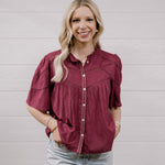 A woman with blonde hair wearing a short-sleeved maroon button-up blouse with decorative stitching and light-wash jeans, standing against a white paneled wall.