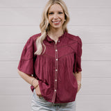 A woman with blonde hair wearing a short-sleeved maroon button-up blouse with decorative stitching and light-wash jeans, standing against a white paneled wall.