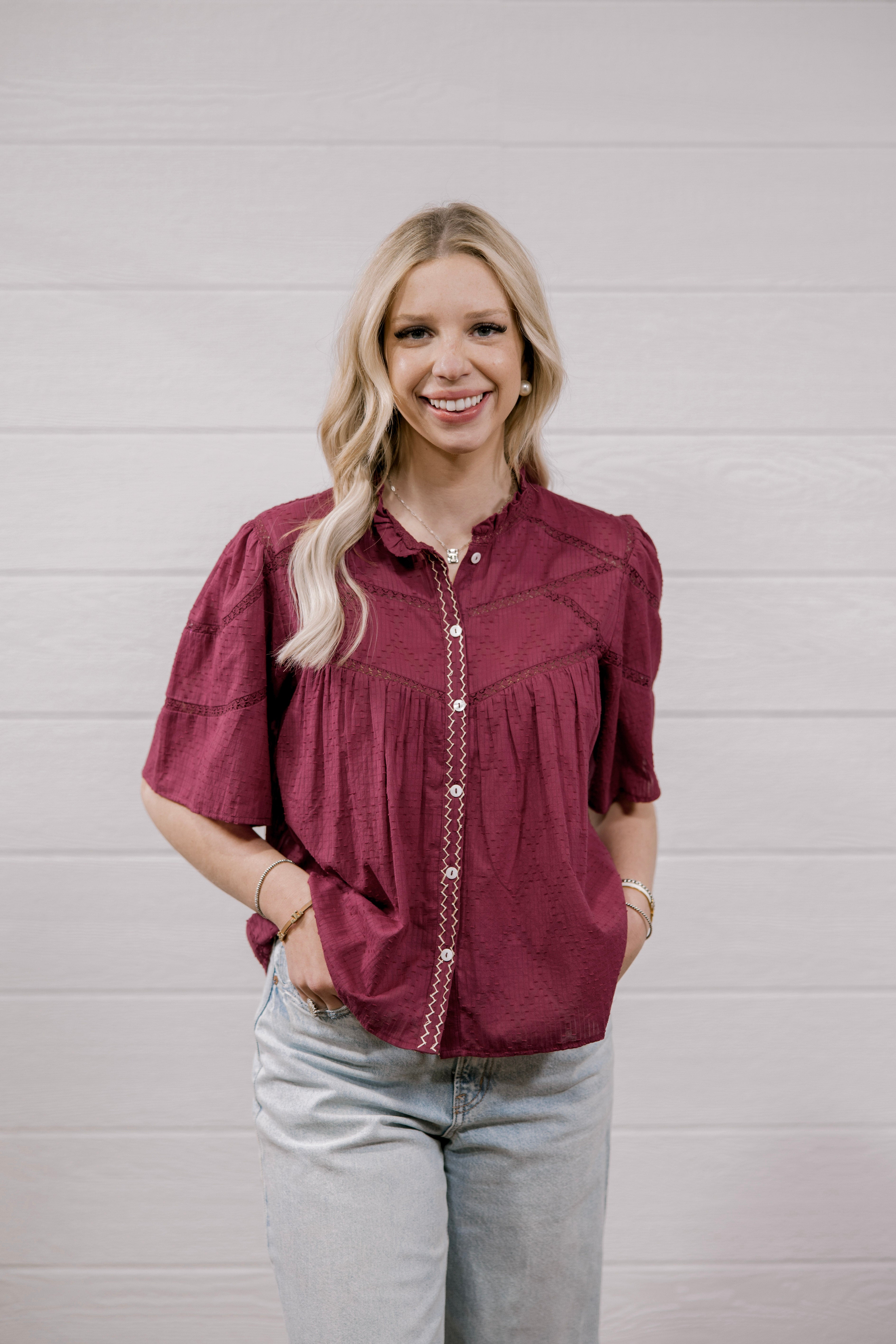 A woman with blonde hair wearing a short-sleeved maroon button-up blouse with decorative stitching and light-wash jeans, standing against a white paneled wall.