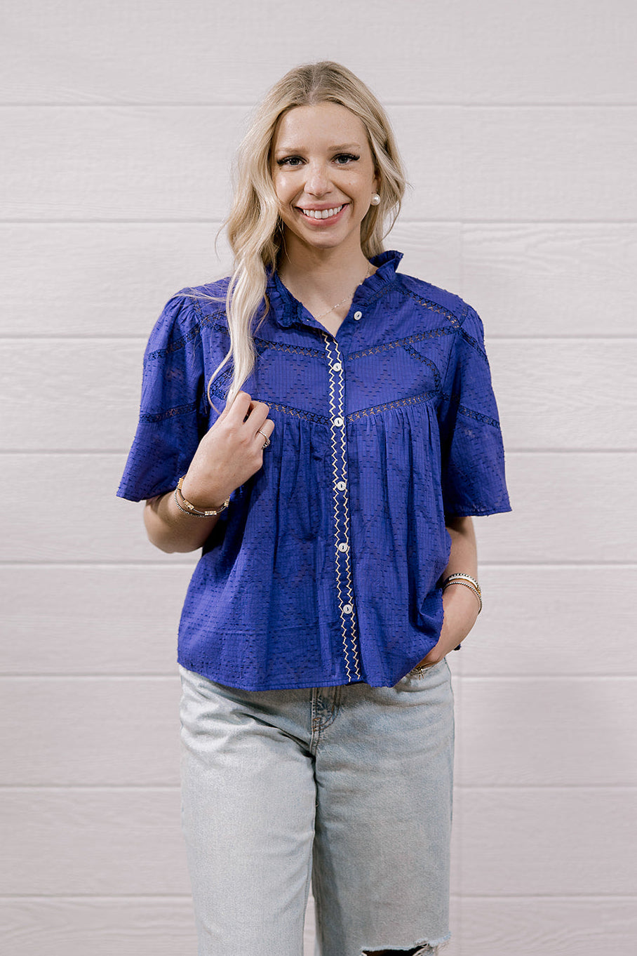 A woman modeling a purple short-sleeve button-up blouse with decorative stitching and light-wash distressed jeans, standing against a white wood-paneled wall.