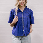 A woman modeling a purple short-sleeve button-up blouse with decorative stitching and light-wash distressed jeans, standing against a white wood-paneled wall.