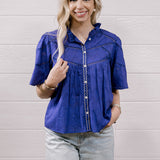 A woman modeling a purple short-sleeve button-up blouse with decorative stitching and light-wash distressed jeans, standing against a white wood-paneled wall.