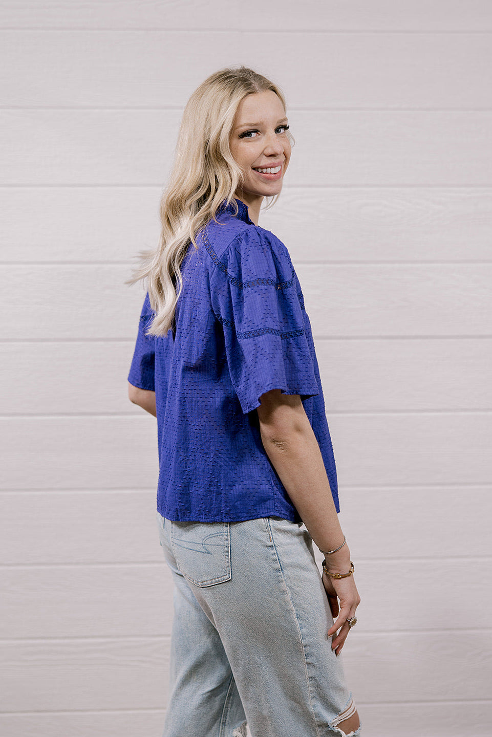 A woman with blonde hair is shown from the back wearing a textured purple short-sleeve shirt and light-wash jeans, standing against a white shiplap wall.