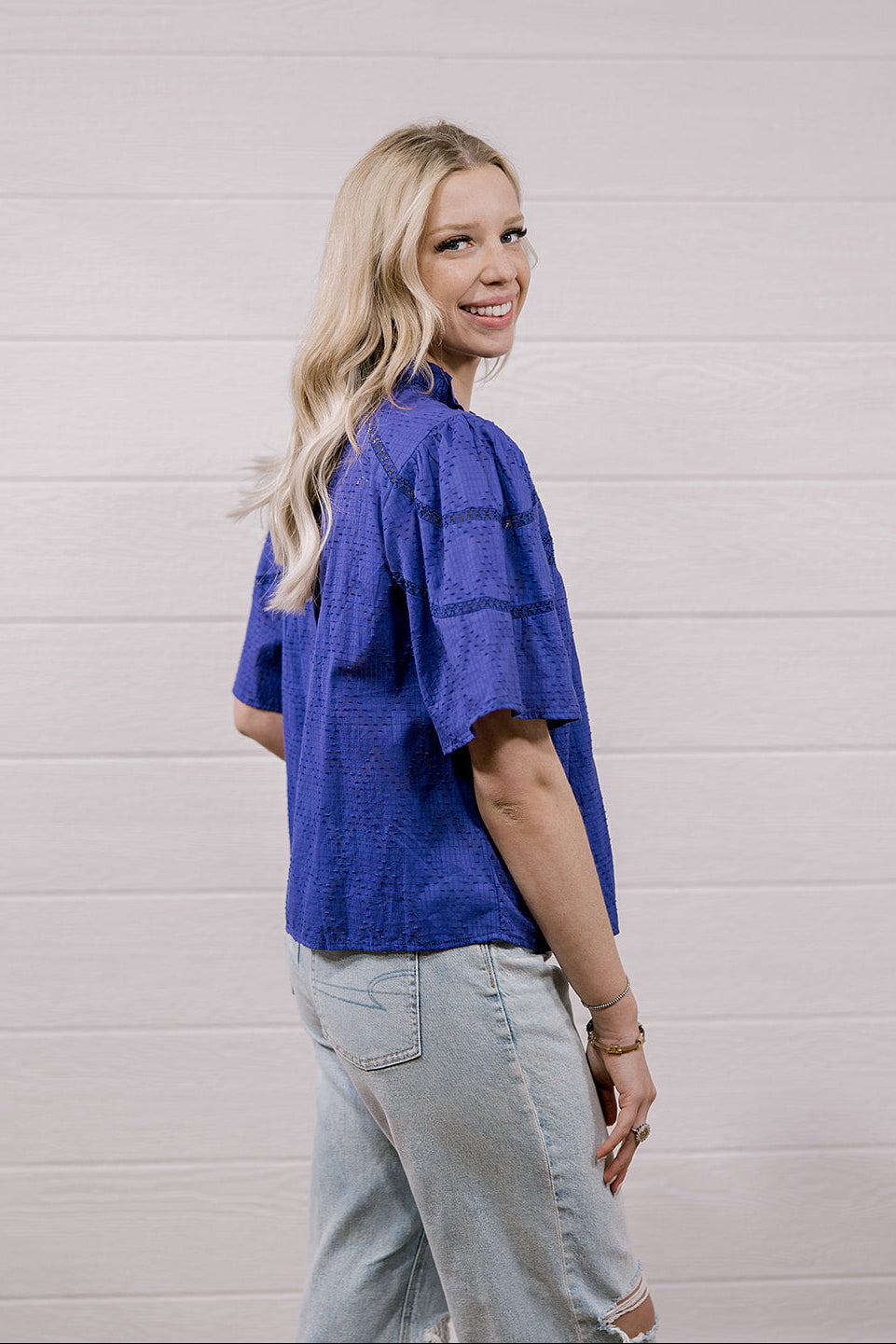 A woman with blonde hair is shown from the back wearing a textured purple short-sleeve shirt and light-wash jeans, standing against a white shiplap wall.