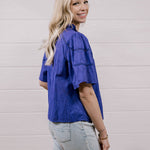 A woman with blonde hair is shown from the back wearing a textured purple short-sleeve shirt and light-wash jeans, standing against a white shiplap wall.